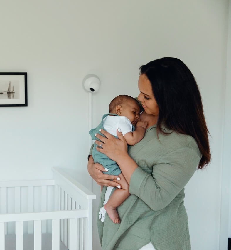 Mother holding baby next to a crib with an Aura baby monitor on the wall